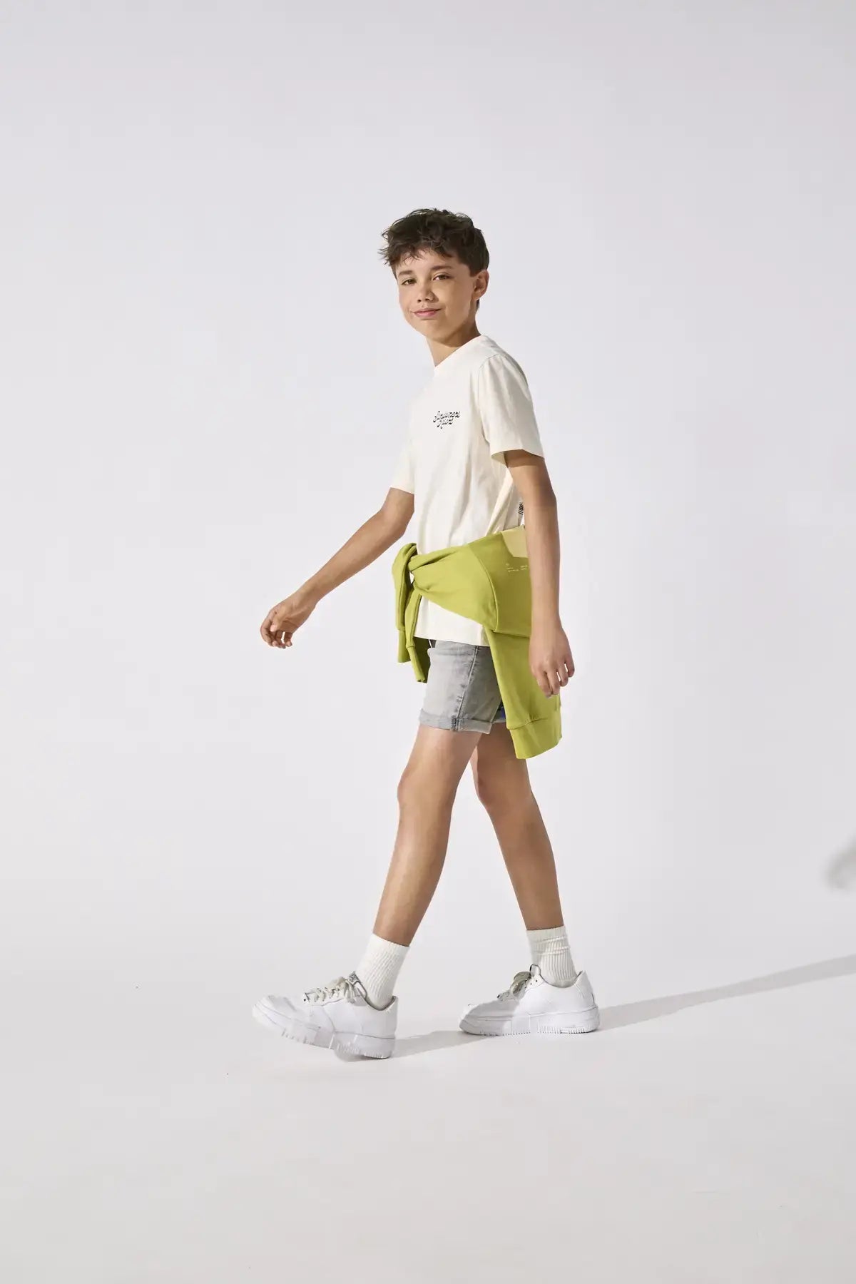 Stylish boy wearing Kids Only & Sons JUNO off white shirt with shorts and a green sweater tied around his waist.